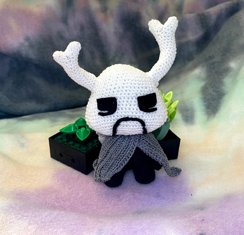 Crochet grumpy critter in a gray cloak in front of two small plants.