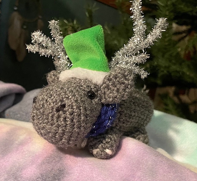 A gray crochet hippopotamus wearing a green Santa Claus hat with sparkling silver pipe-cleaner antlers and a blue scarf. 